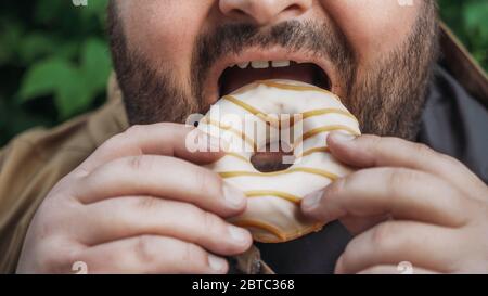 L'uomo grasso mangia ciambella sulla strada, primo piano. Cibi dolci e veloci non salutari. Foto Stock