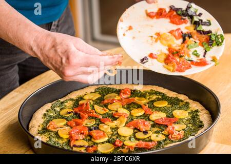 Donna che mette le verdure saltate sulla pizza a grani multipli con salsa pesto Foto Stock