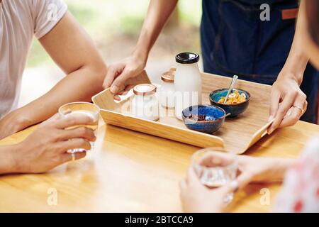 Immagine ravvicinata del cameriere che mette a tavola un vassoio di legno con yogurt e fiocchi di mais davanti alla giovane coppia Foto Stock