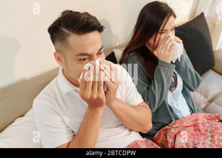 Giovane coppia asiatica malata seduta in letto sotto coperta e soffia nasi in tessuti a casa Foto Stock