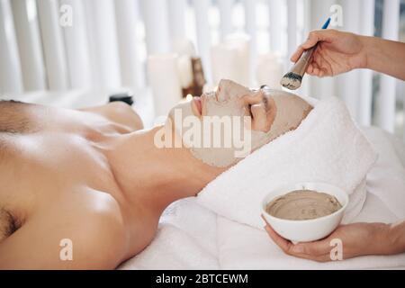 Mani di beautifcian applicando maschera di fango su faccia di giovane uomo che si rilassa in salone di bellezza Foto Stock