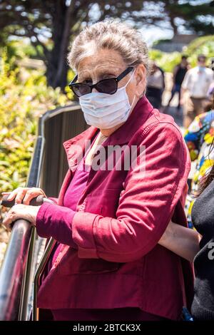 Una donna anziana, 83 anni, indossa una maschera, in piedi da una recinzione, mentre in una passeggiata a Laguna Beach, California, USA. Foto Stock