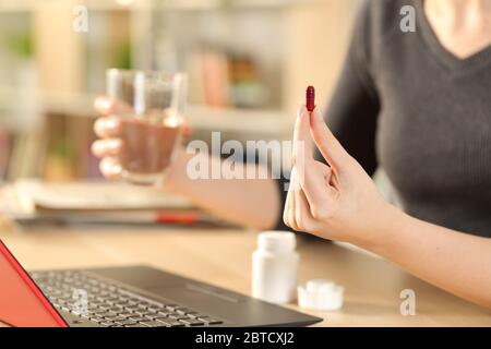 Primo piano di mani di donna che prendono la pillola tenendo il vetro dell'acqua seduto su una scrivania a casa Foto Stock