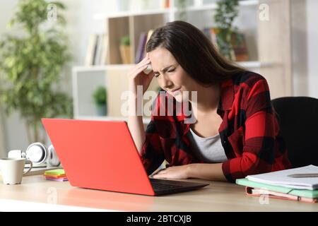 Studentessa con un computer portatile rosso in dolore che soffre di mal di testa seduta su una scrivania a casa Foto Stock