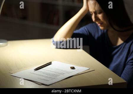 La moglie triste si lamenta dopo aver firmato le carte di divorzio e l'anello di nozze sopra la tabella che siede a casa di notte Foto Stock