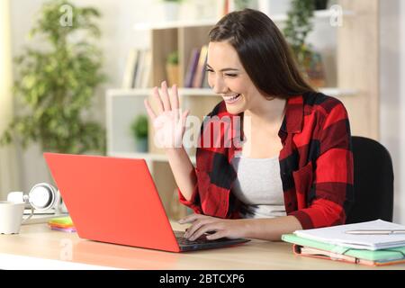 Felice studentessa saluto su videocall su un portatile rosso seduto su una scrivania a casa Foto Stock