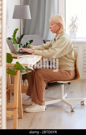 Donna matura che si siede sul posto di lavoro a casa e lavora su un computer portatile Foto Stock