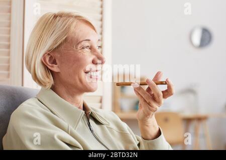 Donna matura sorridente che tiene il telefono cellulare e registra un messaggio mentre si siede sul divano a casa Foto Stock
