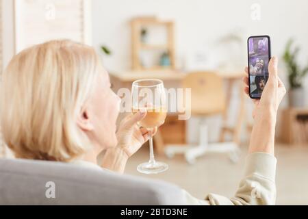 Vista posteriore di una donna matura con bionda capelli corti bere vino e parlare online con la sua famiglia utilizzando il suo cellulare Foto Stock