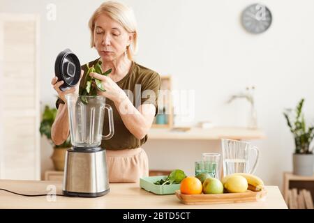 Donna matura che fa un cocktail sano da spinaci e frutta utilizzando il frullatore mentre si trova in cucina Foto Stock