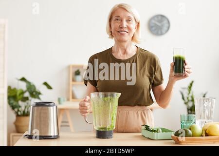 Ritratto di donna matura con capelli biondi sorridenti alla macchina fotografica e bere succo di verdure fresco mentre si trova in cucina Foto Stock