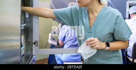 Una dottoressa tiene in mano una maschera medica e toglie le forniture mediche da un armadietto. Un medico in uniforme verde. In background sono docto Foto Stock