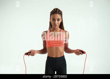 Sicuro e sano. Primo piano di una giovane donna africana che tiene la corda saltante mentre si trova in studio su sfondo grigio. Attrezzature sportive. Stile di vita attivo Foto Stock