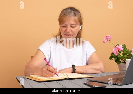 Donna anziana sorridente che indossa le cuffie ascoltando il corso audio utilizzando il computer portatile a casa, facendo appunti, femmina di mezza età imparare le lingue straniere Foto Stock