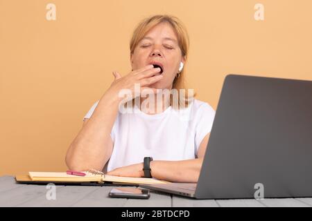Donna anziana di mezza età seduta al tavolo a casa lavorando con computer portatile annoiato sbadigli stanchi coprendo la bocca con la mano. Foto Stock
