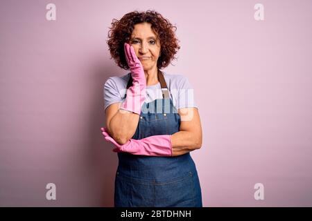 Media età capelli ricci donna pulizia facendo lavoro domestico indossare grembiule e guanti pensando stanco e annoiato con problemi di depressione con attraversato A. Foto Stock