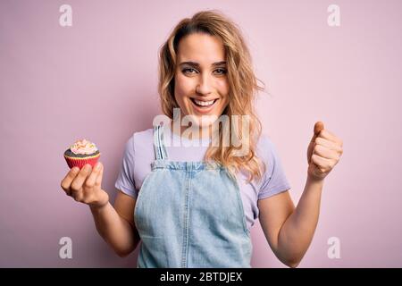 Giovane bella donna bionda eatimg cioccolato cupcake su isolato sfondo rosa urlando orgoglioso e celebrando la vittoria e il successo molto eccitato, Foto Stock