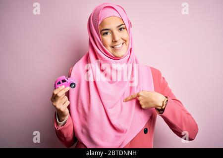 Giovane ragazza che indossa hijab musulmano tenendo un piccolo giocattolo per auto su sfondo rosa isolato con faccia a sorpresa che punta il dito verso se stesso Foto Stock