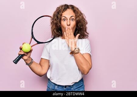 Media età bella sportivo che gioca a tennis tenendo racchetta e palla su sfondo bianco che copre la bocca con la mano, scioccato e impaurito per errore Foto Stock