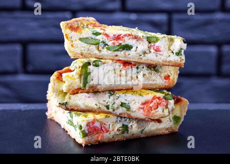 primo piano di una pila di fette di quiche con asparagi di salmone ripieno su un tavolo di cemento con un muro di mattoni sullo sfondo, vista orizzontale Foto Stock