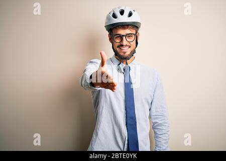 Giovane uomo d'affari che indossa occhiali e casco da bici in piedi su isolato panettaio bianco sorridente amichevole offrendo handshake come saluto e accogliente Foto Stock