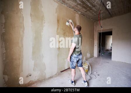 Vista posteriore di un ragazzo che tiene in mano lo spray di vernice e dipingere un muro di giorno. Il maschio è vestito con indumenti ricoperti di vernice. Ristrutturazione della casa. Foto Stock