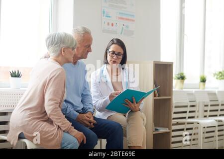 Ritratto di giovane dottore che parla con la coppia anziana mentre si siede nell'area di ricevimento della clinica moderna, spazio di copia Foto Stock
