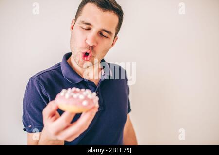 Giovane isolato su sfondo bianco. Il ragazzo emozionale tiene in mano una ciambella gustosa e la guarda. Andare a mangiare. Deliziosa ciambella fatante Foto Stock