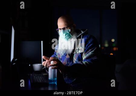 Uomo con palma mature con maschera usando igienizzatore per le mani mentre si lavora da casa al buio Foto Stock