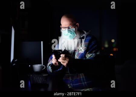 Uomo calvo maturo con maschera utilizzando il telefono mentre si lavora da casa a tarda notte Foto Stock