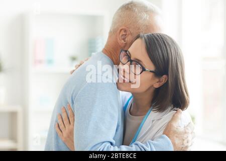 Vita in su ritratto di giovane dottore femmina abbracciando paziente senior grato e sorridendo felice mentre si trova in ufficio, copia spazio Foto Stock