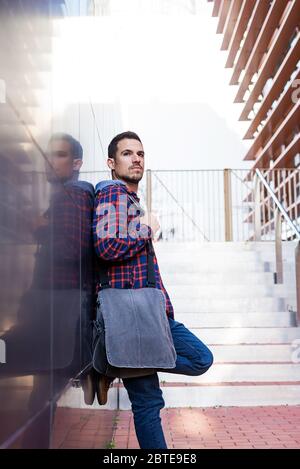 Ragazzo con il cuscinetto poggiato su una parete scura che tiene una borsa a spalla mentre si guarda via Foto Stock