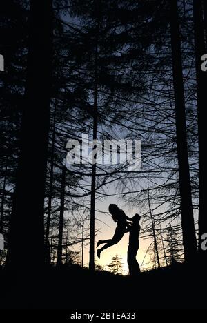 Da sotto si può vedere le silhouette di coppia amorevole che si posano nella foresta serale. Natura selvaggia con viste incredibili, tramonto estivo, uomo che tiene la donna mentre salta e si sente felice. Natura, concetto di amore. Foto Stock