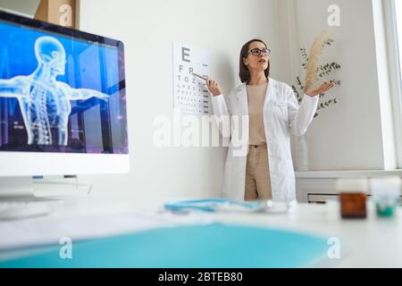 Ritratto grandangolare di giovane oftalmologo femminile che punta alla mappa oculare durante il test di vista in clinica, spazio di copia Foto Stock
