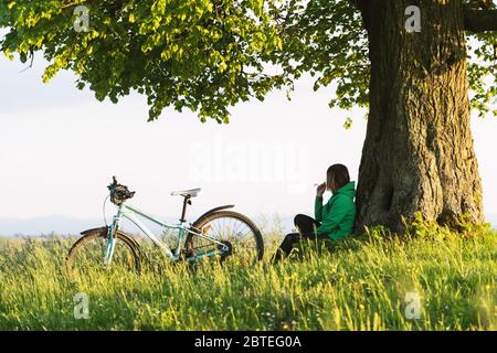 Una ragazza sola si siede sotto un grande vecchio albero vicino ad una bicicletta. Verde paesaggio estivo sul lato opposto della città Foto Stock