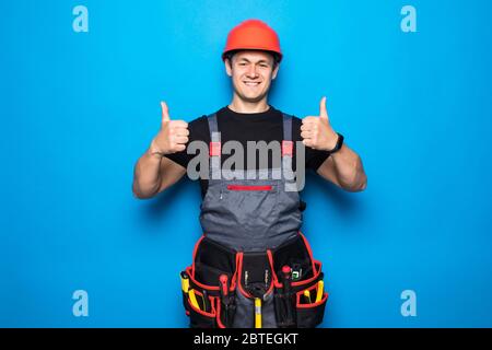 Felice giovane lavoratore sorridente e pollici, ragazzo indossando indumenti da lavoro e cintura attrezzature e hemlet isolato su sfondo giallo Foto Stock