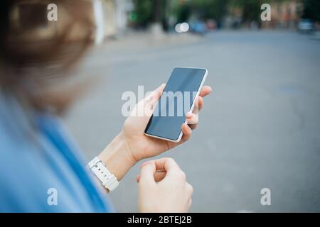 Telefono cellulare con schermo vuoto in mano di giovane donna che cammina lungo la strada della città di giorno, primo piano. Foto Stock