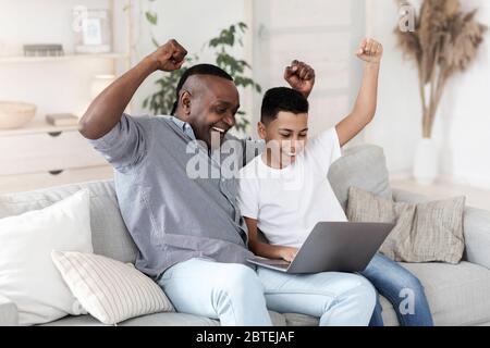 Nonno nero gioioso e nipote che guardano gli sport sul portatile e che emozionano Foto Stock