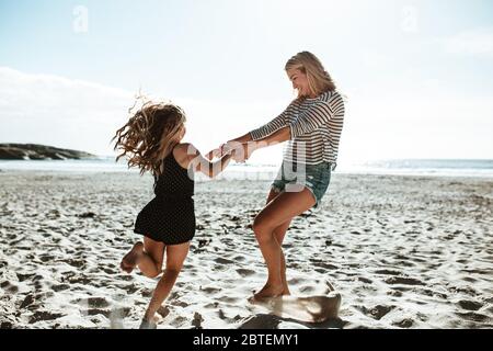 Donna sorridente che gioca con una ragazza sulla spiaggia. Madre e figlia che tengono le mani che giocano sulla riva.