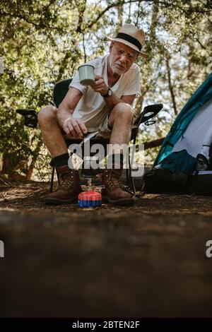 Uomo anziano che fa il caffè all'aperto in un viaggio in campeggio. Uomo maturo seduto fuori della tenda al campeggio preparazione del caffè sulla stufa del campo. Foto Stock