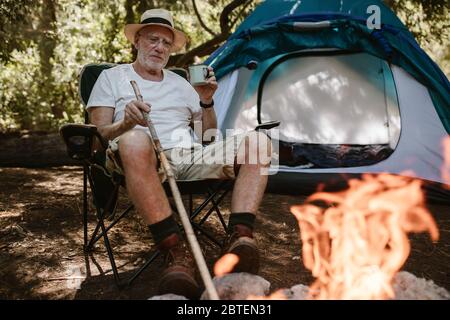 Uomo in pensione seduto rilassato vicino al falò e con una tazza di caffè. Uomo anziano che si accampano in foresta con un caffè rinfrescante al mattino. Foto Stock