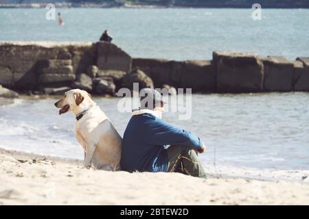 L'anziano e il suo cane seduti di nuovo su una piccola spiaggia a Lisbona, in Portogallo, l'uomo che guarda al mare, assomiglia a un pescatore Foto Stock