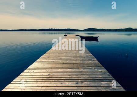 Una barca di legno con remi ormeggiati ad un molo su un lago in primavera. Il molo dalle tavole parte con la prospettiva verso l'orizzonte. Il sole nel bl Foto Stock