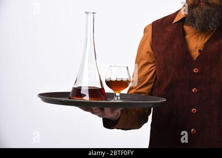 Il barman in pelle scamosciata vintage è un gilet che serve scotch o brandy. Cameriere con bicchiere e bottiglia di whisky sul vassoio. Concetto di bevande del ristorante. L'uomo con la barba tiene il cognac su sfondo bianco. Foto Stock