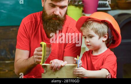 Padre, genitore con barba insegnando il bambino a segare mentre il figlio gioca con la sega giocattolo. Concetto di giochi educativi. Ragazzo, bambino occupato in casco protettivo imparare ad usare la sega a mano con papà. Foto Stock