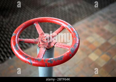 Vecchia ruota di valvola arrugginita vernice rossa verniciata con rigonfiamenti e peeling fuoco selettivo con dispositivo di estensione su sfondo fuori fuoco pavimento piastrellato e. Foto Stock