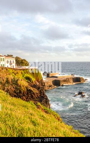 Scogliere sull'oceano Atlantico nel villaggio di pescatori Lagoa, Sao Miguel Island, Azzorre, Portogallo. Case tradizionali sulla collina sopra il mare. Luce del tramonto. Foto verticale. Foto Stock