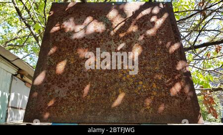 Vecchia superficie metallica arrugginita marrone con macchie sfocate di luce solare e ombra. Lamiera trapezoidale di metallo corroso con sfondo verde sfocato Foto Stock