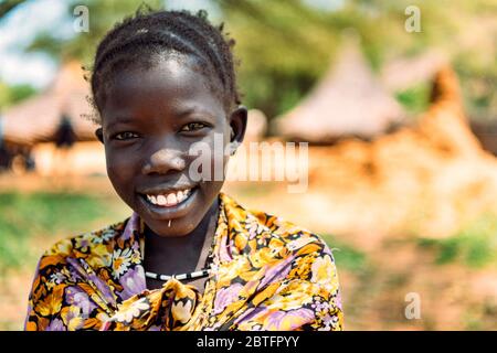 TRIBÙ BOYA, SUD SUDAN - 10 MARZO 2020: Ragazza in abiti colorati e accessori tradizionali sorridendo a macchina fotografica contro insediamento offuscato nel Sud Foto Stock