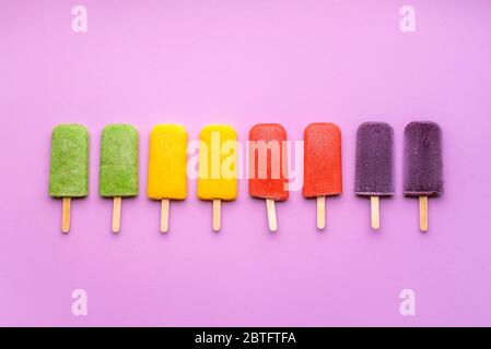 Assortimento di papsicle di gelato alla frutta su sfondo viola. Piatto con gelato arcobaleno su bastone line up. Dessert estivo rinfrescante. Sorbetto vegano Foto Stock